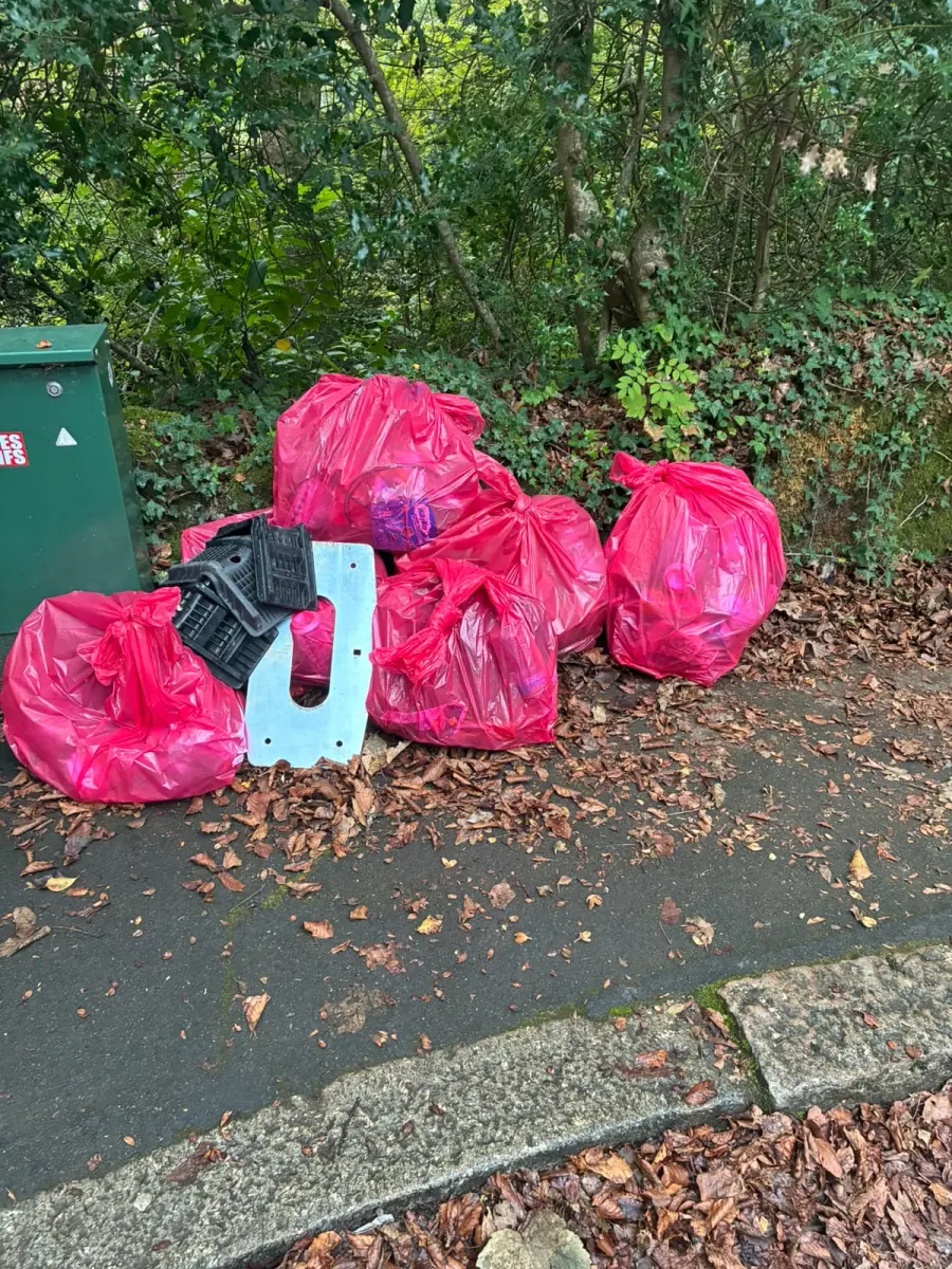 Six tied pink sacks of litter piled on the pavement beside a green cabinet, with broken black plastic pieces and a galvanised steel plate on top