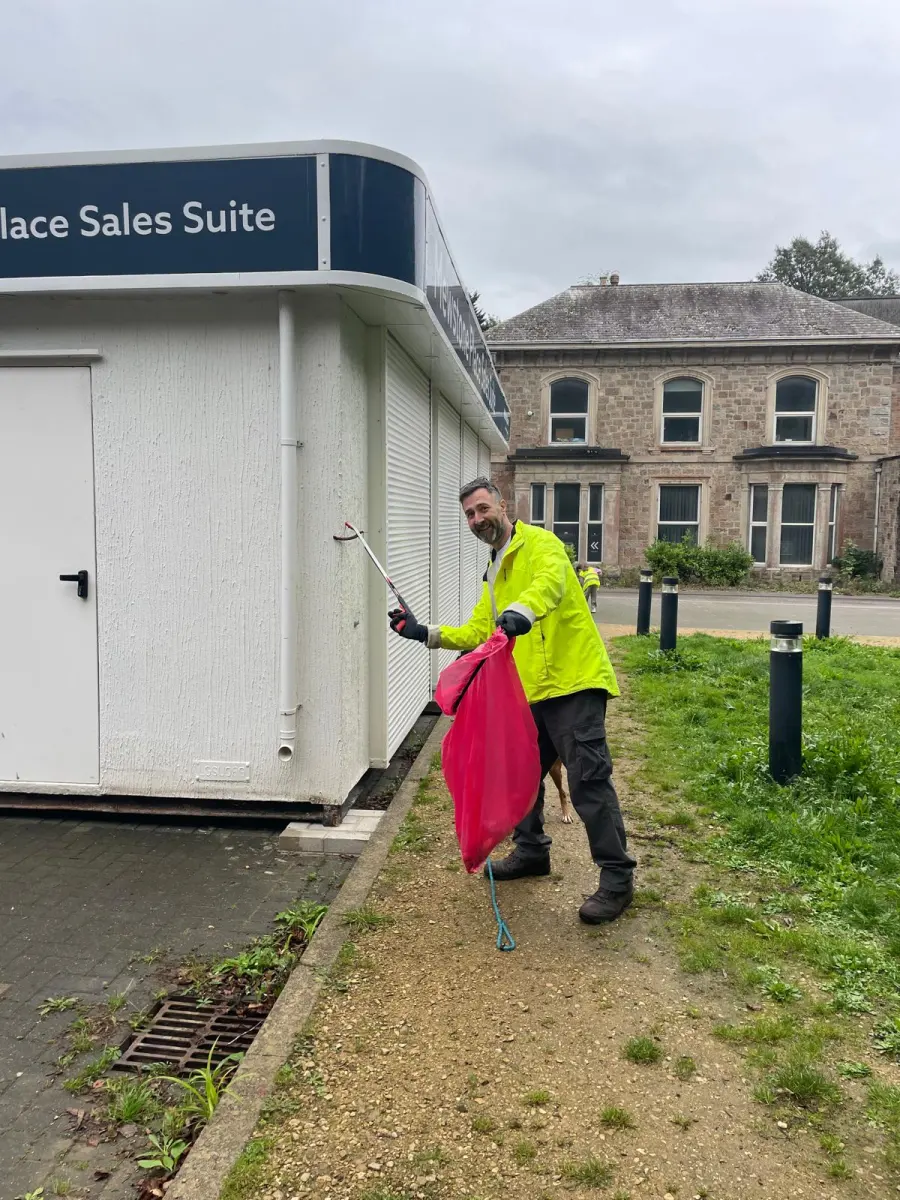 A resident in a high-vis jacket picking litter beside the sales suite, a pink sack in one hand and a dog on a lead