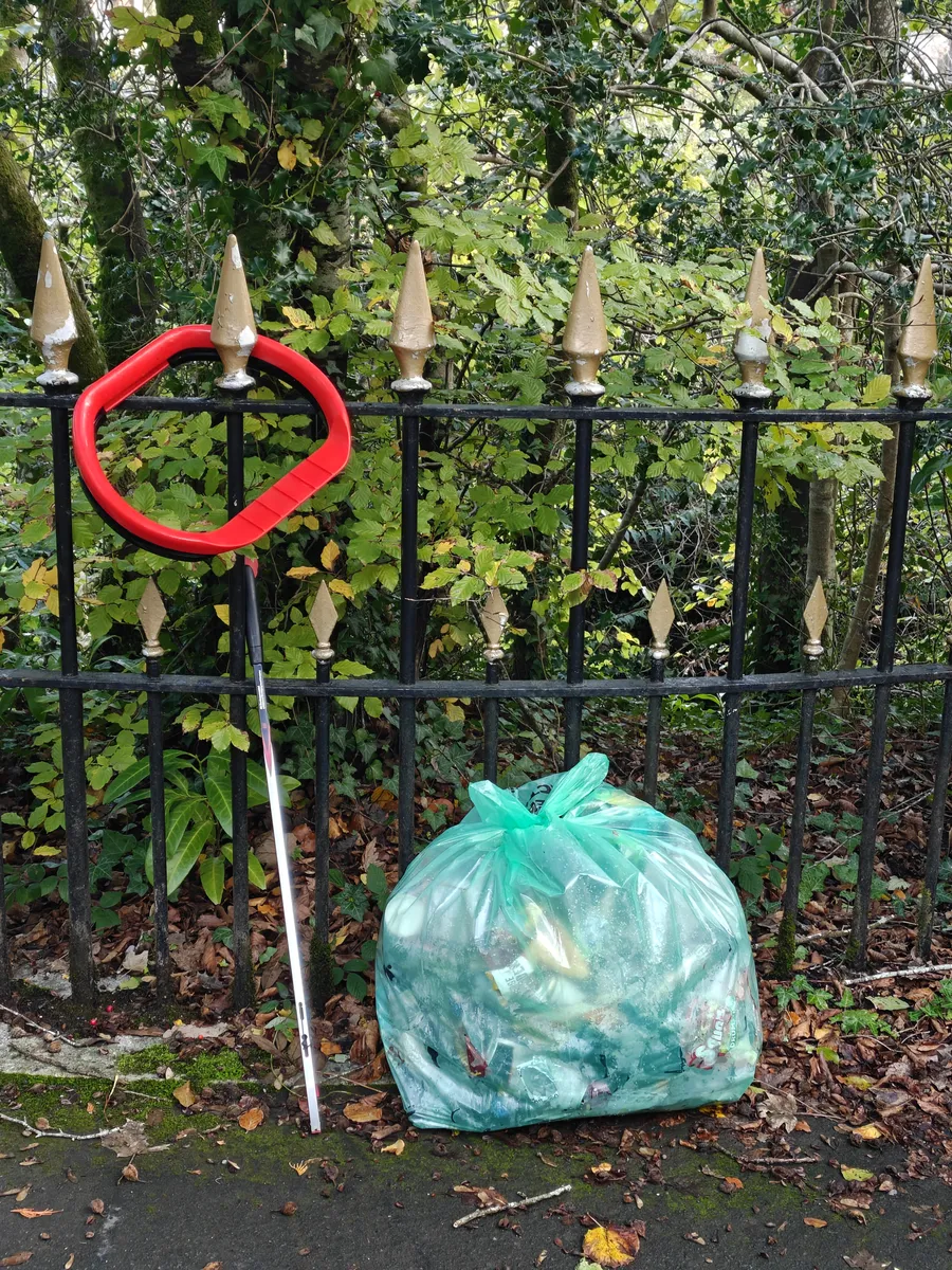 A full, tied green sack of litter on the pavement by black iron railings, with a litter picker and a red litter hoop hung on a railing spike
