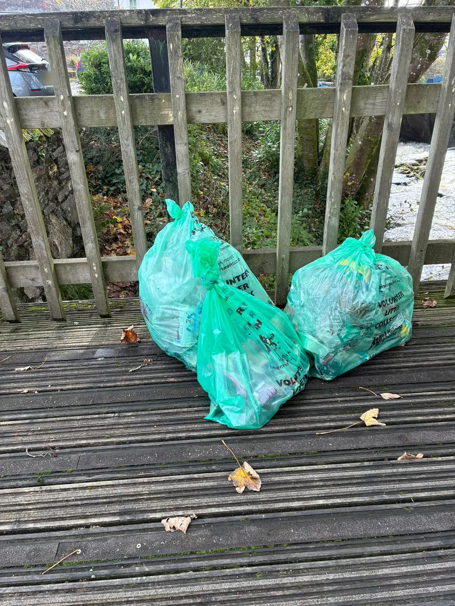 Three full, tied green volunteer litter sacks on the decking of a wooden footbridge over the river