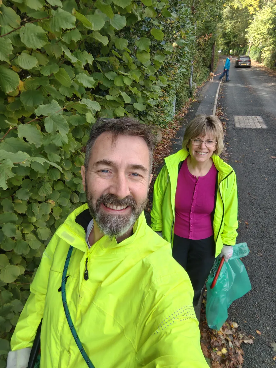 Nat and Jan in high-vis jackets smiling on a tree-lined road, Jan holding a green sack and a litter picker